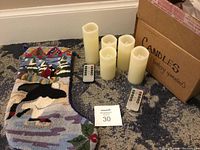 Nine cream colored battery operated candles of varying heights from about 4 inches to 8 inches, two remote controls with buttons, standing next to a cardboard box and a penguin design hook stocking with needlepoint style and velvet back.