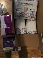 Photo showing unopened bags of Because Premium Maximum bladder control underwear and Poise pads partially unpacked from a box on the floor.