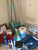 Photo showing multiple Swiffer mops, broom, dust pan, cleaning sprays in red, blue and green bottles, and paper towels arranged together.