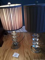 Two matching acrylic table lamps with stacked diamond-shaped bases and taupe pleated fabric shades shown on floor.
