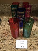 Five vintage colorful drinking glasses in red, green, blue, and pink with gold ornate designs shown on a kitchen counter with auction card.