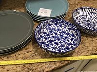 Four grey plates, four blue plates, and two blue and white patterned serving bowls on kitchen counter with measuring tape for scale.