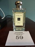 Front view of Jo Malone Pomegranate Noir Cologne bottle showing label and cap, placed on a dark wooden table with auction tag in front.