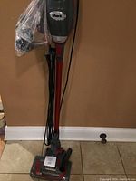 Full view of Shark Rocket upright vacuum cleaner with power cord wrapped around it and attachments in plastic bag.