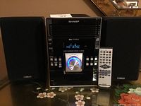 Front view of Sharp micro component system flanked by two black Micca speakers and white remote placed beside the system, all on a floral tablecloth.