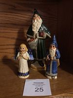 Set of three Vaillancourt Folk Art figurines displayed on a wooden shelf: two Father Christmas and one angel figurine.