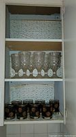 Shelf with six clear cut glass water goblets on upper shelf and eight black bicentennial rocks glasses with gold medallions on lower shelf.