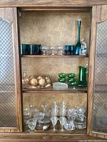 Full view of wooden cabinet with three shelves filled with various glassware including blue decanter, green pitcher, gold decanter with stemware and shot glasses, and clear glass dishes and stemware.