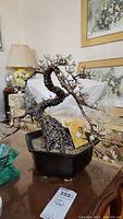 Front angled view of the metal bonsai tree sculpture showing textured trunk, branches with white and yellow metal blossoms, and black planter pot with gravel.