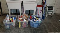 Wide view of basement floor with laundry tub, three bins filled with assorted cleaning and laundry supplies, and two white step stools.