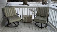 Two outdoor swivel chairs with green and beige striped cushions on a porch, with a small matching side table covered with fabric between them.