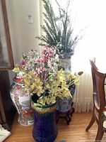 Three decorative ceramic vases shown grouped together, one with multi-colored faux flowers, the other with tall green faux foliage, one white vase with a wood base.