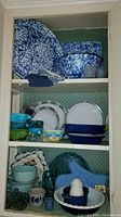 Photo showing full cabinet view with enamelware on top shelf, mixed bowls and plates on middle shelf, and lower shelf with additional kitchen items including small cups.