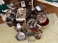 Wide shot showing a variety of silverplate items including platters, bowls, coffee and tea pots, and more arranged on and around small tables.