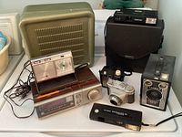 Group photo of vintage cameras, clock radios, telephone handset, and cords on a surface.