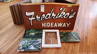 Photo of wooden sign with 'Fredriko's Hideaway' text and three nature-themed canvas prints plus a framed glass tree painting.