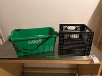 Photo showing the green plastic grocery basket and black plastic milk crate side by side on metal table, labeled with lot sticker.
