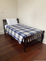 Twin bed frame with blue plaid bedspread, white pillow and sheet against a wall on hardwood floor.