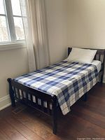 Full view of the twin bed frame against a wall beneath a window, made of dark hardwood with bed linens and pillow.