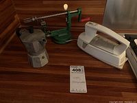 Bialetti espresso pot, GE hand mixer, and manual apple peeler displayed on a wooden surface.