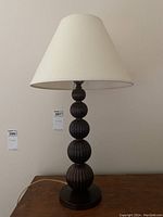 Metal table lamp with five stacked ribbed spheres as the base and a fabric shade with indentation marks, shown unlit against a neutral background.