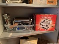 Imperia pasta maker machine, additional pasta cutter attachment, clamp, wooden handle crank inside a plastic container alongside the original product box on a shelf.