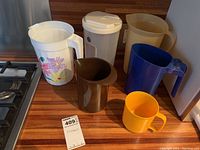 Photo showing all 5 plastic pitchers and 1 yellow plastic cup on a wooden counter near a stove.