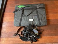 Top view of black nylon portfolio case with handles and front zippered pocket, laid flat on wood floor with extra bag straps beside it.