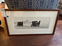 Framed black and white print of winter buggies with horses, shown standing upright on the floor with couch in background