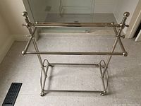 Front view of the entire silver metal towel rack with two horizontal bars and lower support on a tiled floor.