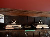 Two rectangular silver-tone serving dishes with lids and decorative handles alongside a round silver-tone plate with clear glass insert and two spoons