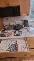Full view of kitchen counter and open drawer showing Corningware dish, ceramic bowls, platters, box of Resinite foil, and tea towels in drawer.