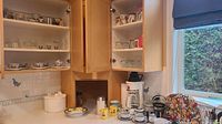 Kitchen cabinet with shelves holding various stoneware cups, glassware, coffee maker and accessories, yellow mugs, and small decorative bowls.