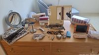 Wide view of the lot on a wooden dresser including mirrors, costume jewelry, curling irons, digital picture frame, rollers, and scalp massager, shown for overall content and item variety.