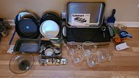 Full view of the lot items arranged on a kitchen counter including Cuisinart electric grill and griddle, metal bakeware, glass mixing bowls, measuring spoons, and kitchen utensils.