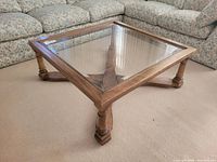 Square coffee table showing wood base with turned legs and clear glass top in place, positioned on carpeted floor with floral upholstered couches surrounding.