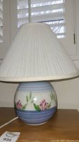 Ceramic table lamp with hand painted pink flowers and green leaves on a blue band base topped with a white fabric pleated lampshade.