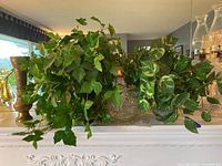 Two artificial plants displayed on a white mantle, one in a clear crystal-like glass vase and the other in a brass-colored metal vase.