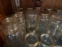 Multiple mostly quart size wide mouth canning jars on wooden shelf. Some have metal lids, some without lids. Glass is clear, jars have varying heights and some show embossed designs.