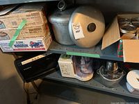 Shelf with kitchen appliances and canning supplies including pressure cooker, jars, Food Saver vacuum sealer, Braun food processor, metal bowls.