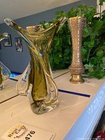 Photo showing chalice glass vase on white piano with some other decorative metal vase in the background