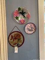 Wall display of three decorative items: glass hummingbird art, floral plate, and a welcome sign; focus on two primary art pieces.
