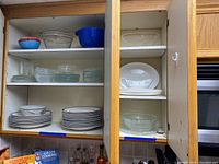 Open kitchen cabinet showing stacks of 20 white ceramic dinner plates, clear glass dessert dishes and platters in various sizes and shapes.