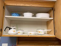 Vintage white ceramic Corning Ware pots and dishes with floral pattern, including a larger casserole dish and smaller pots, displayed in wooden cabinet.