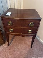 Front view of the wooden end table showing two drawers with oval brass handles, curved legs, and polished top surface.