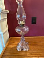 Clear glass hurricane oil lamp with ornate patterned base and chimney standing on wooden surface against red wall.