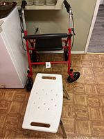 Red walker and white shower chair shown in the same photo, walker positioned behind shower chair in a room with linoleum and a white appliance on the left