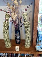 Two tall pale green resin figurines flanking a black floral peacock vase on wooden shelf