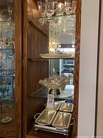 Overview of the glass cabinet shelf showing assorted glassware, a cut glass bowl, and a rectangular divided silver-tone serving tray with lid.