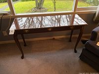 Full view of the wooden console table placed against a window, showing the polished top and the three faux drawer fronts with decorative handles.